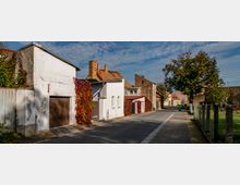 Straßenszene in einem Dorf mit gepflastertem Gehweg, kleinen Häusern in weißer und roter Ziegeloptik und wildem Efeu an den Fassaden. Rechts verläuft ein Zaun entlang einer grünen Wiese, im Hintergrund stehen Bäume und weitere Gebäude.