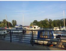 Yachthafen mit mehreren Segel- und Motorbooten, die an Stegen festgemacht sind. Im Hintergrund Bäume und eine ruhige Wasserfläche unter klarem Himmel, vorne ein Geländer entlang der Uferpromenade.