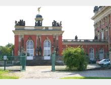 Historisches Gebäude mit roten Ziegelwänden, weißen Rundbogenfenstern und einer verzierten Balustrade mit Figurenstatuen. Vor dem Gebäude befindet sich ein schmiedeeisernes Tor sowie eine gepflasterte Fläche, umgeben von Grünflächen.
