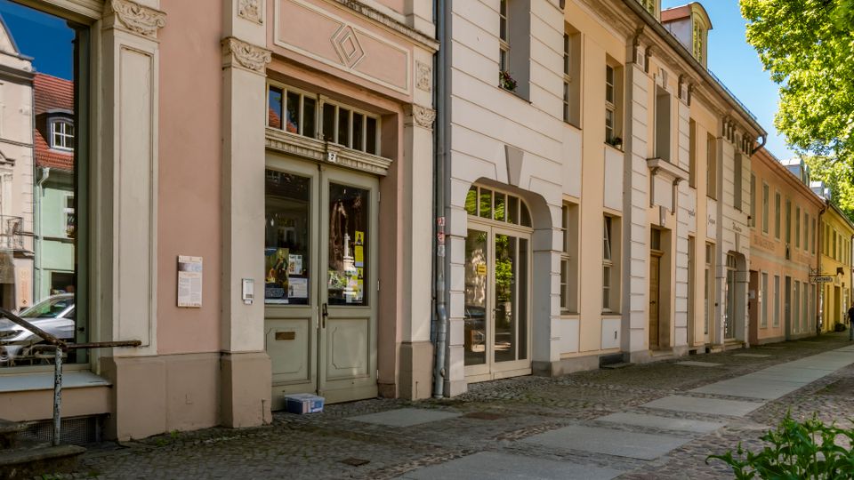 Straßenzug mit historischen, pastellfarbenen Gebäudefassaden mit hohen Fenstern und verzierten Türrahmen, teils mit Ladeneingängen im Erdgeschoss. Breiter Gehweg mit Kopfsteinpflaster und großen Straßenbäumen entlang der rechten Seite.