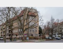 Eckgebäude eines mehrstöckigen Wohnhauses mit gelblich-grauer Fassade und rotem Satteldach, umgeben von kahlen Bäumen. Im Vordergrund eine Straßenkreuzung mit Fußgängern, Autos und einer Ampel.