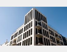 Modernes Mehrfamilienhaus mit weißer Fassade, großen vertikalen Fenstern und mehreren Balkonen. Das Gebäude hat eine klare Linienführung und eine Dachterrasse mit Glasgeländer unter einem wolkenlosen Himmel.