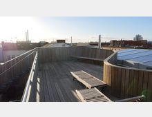 Holzterrasse auf einem Flachdach mit geschwungener Holzverkleidung und integrierten Sitzbänken, ergänzt durch zwei liegende Holzliegen. Im Hintergrund sind umliegende Dächer und Schornsteine einer Stadtlandschaft bei Tageslicht zu sehen.