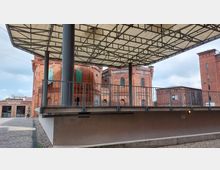 Außenbereich mit Blick auf ein historisches Backsteingebäude mit runden und rechteckigen Fenstern, teilweise mit grünen Fensterläden. Im Vordergrund eine große, moderne Überdachung mit Metallstreben und Geländer, sowie eine Pflasterstraße und ein ausgeschilderter Eingang zur Rezeption auf der linken Seite.