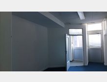 Leerer Raum mit weißen Wänden und blauem Teppichboden. Im Vordergrund eine geöffnete Tür, die zu einem Bereich mit Fenstern und einer Glastür führt; darüber sind Oberlichter sichtbar.