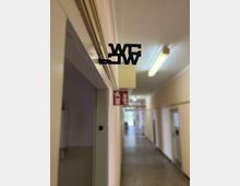 Langer Innenflur eines Gebäudes mit mehreren Türen entlang der Wände, grauem Boden und Deckenleuchten. Im Vordergrund hängt über einem Türrahmen ein schwarzes „WC“-Schild; weiter hinten ist ein rotes Brandschutzschild zu sehen.