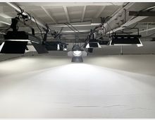 Eine Studioaufnahme mit einer weißen Wand und mehreren professionellen Scheinwerfern sowie Studiolichtern, die von der Decke hängen. Deutlich sichtbar sind Kabel und Klemmen an den Metallgerüsten, die die Beleuchtung tragen.