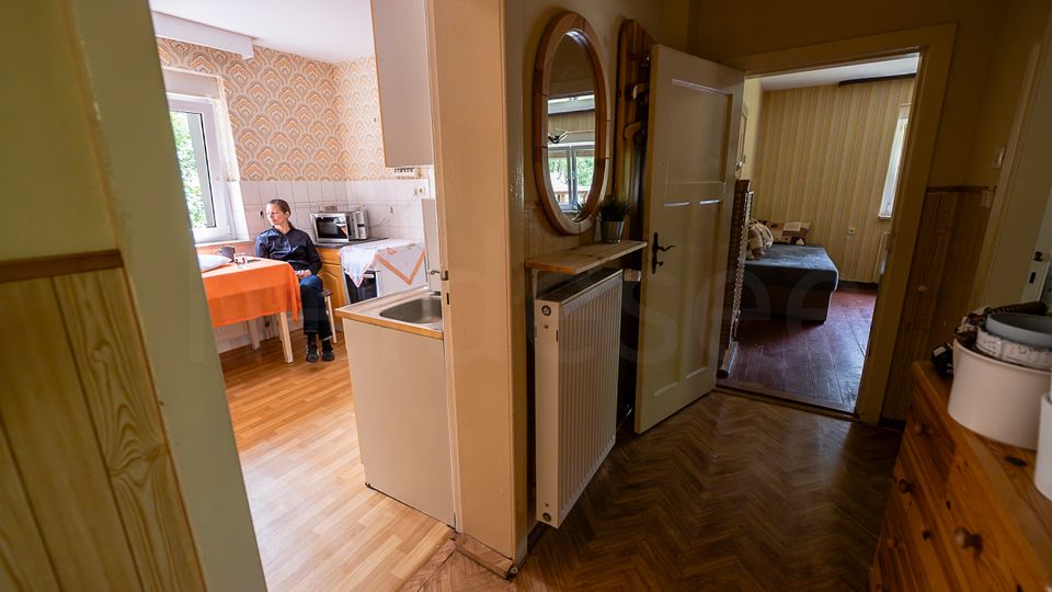Blick durch einen Flur in eine ältere Wohnung: links eine kleine Küche mit gemusterter Tapete, Fenster und Esstisch, rechts ein schmaler Gang mit Holzfußboden, Rundspiegel und Heizkörper. Im Hintergrund führt eine offene Tür in ein Schlafzimmer mit Bett und gestreifter Tapete.