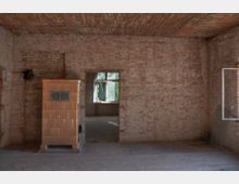 Ein leerer Raum mit unverputzten Ziegelwänden und einer Holzdecke. Links steht ein hellbrauner, gefliester Ofen, und in der Mitte führt eine Türöffnung zu einem benachbarten Raum mit einem Fenster, durch das Tageslicht einfällt.