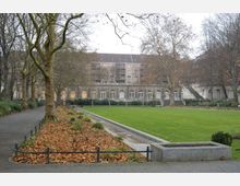 Ein gepflegter Park mit einer großen Rasenfläche, gesäumt von Laubbäumen und einem gepflasterten Weg. Im Hintergrund steht ein historisches Gebäude mit großen Fenstern und dekorativen Steinbalustraden, dahinter mehrstöckige Wohnhäuser.