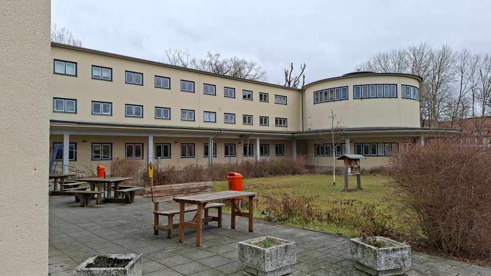 Innenhof einer modernen, hell verputzten Gebäudeanlage mit drei Stockwerken und vielen rechteckigen Fenstern, rechts mit einem abgerundeten Gebäudeteil. Im Vordergrund stehen Holz-Picknicktische auf gepflasterter Terrasse neben einer Rasenfläche mit Sträuchern, Pflanzkübeln und einzelnen Bäumen.