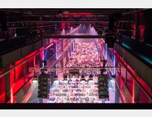 Große Veranstaltungshalle mit vielen Sitzreihen, gefüllt von einer Menschenmenge. Die Halle hat eine hohe Decke, rote und blaue Lichteffekte sowie eine sichtbare Beleuchtungs- und Lautsprecheranlage an der Decke.