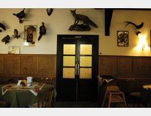 Innenraum eines rustikalen Restaurants mit dunklen Holzmöbeln, grün gedeckten Tischen und einer Holzwandverkleidung. Über der Wand hängen ausgestopfte Tiere und dekorative Bilder, in der Mitte eine Doppeltür mit gelblichen Glaseinsätzen.
