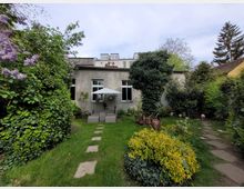Ein kleiner Garten mit grüner Rasenfläche, blühenden Sträuchern und dichtem Laub umgibt ein Haus mit beige verputzter Fassade und großen Fenstern. Im Vordergrund stehen Gartenmöbel mit einem Sonnenschirm, und ein gepflasterter Weg führt durch das dichte Grün rund um das Gebäude.