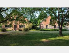 Zwei Backsteingebäude in ländlicher Umgebung, umgeben von Bäumen und einer Wiese. Im Vordergrund werfen die Äste eines großen Baumes Schatten auf die Grünfläche.