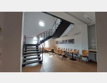 Innenraum eines Gebäudes mit einer Metalltreppe, die in das obere Stockwerk führt. Im unteren Bereich stehen Tische und Stühle auf einem beigen Fliesenboden, an der Wand hängen mehrere Bilder, und durch Rundfenster fällt Tageslicht in den Raum.