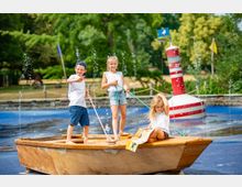 Drei Kinder stehen oder sitzen in einem hölzernen Spielboot in einem Wasser-Spielbereich mit sprudelnden Fontänen. Im Hintergrund sind grüne Bäume und eine rot-weiß gestreifte Boje sichtbar.