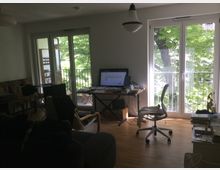 Wohnzimmer mit Holzfußboden, einem Schreibtisch mit Computer am rechten Fenster und einem Drehstuhl davor. Große Fensterfronten auf beiden Seiten lassen viel Tageslicht herein und bieten Blick auf grüne Bäume. Links befindet sich ein Balkon mit Zugang durch eine geöffnete Balkontür.