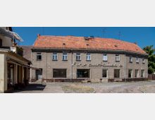 Zweistöckiges Gebäude mit grauer Fassade und rotem Ziegeldach, auf dessen Vorderseite ein Schriftzug mit unleserlichen Buchstaben angebracht ist. Im Vordergrund ein gepflasterter Platz, links daneben ein kleiner Anbau mit Markise.