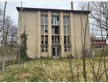 Verlassenes, mehrstöckiges Gebäude mit beigem Putz und großen, teils zerbrochenen Fenstern, umgeben von einer überwucherten Wiese und kahlen Bäumen. Im Hintergrund ist ein weiteres Industriedach zu sehen.