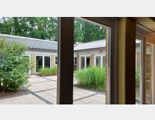 Blick aus einem Fenster auf einen begrünten Innenhof mit gekachelten Wegen, bodentiefen Fenstern an einem Gebäudering aus gelben Ziegeln und einem flachen Metalldach; dahinter eine Baumreihe.