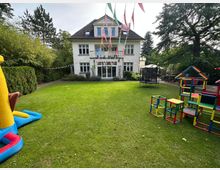 Zweistöckiges Gebäude mit hellen Wänden und dunklem Dach, umgeben von einem Garten mit Rasenfläche. Im Vordergrund stehen Spielgeräte aus bunten Kunststoffstangen, eine Hüpfburg und ein Trampolin, dekoriert mit bunten Wimpelketten.
