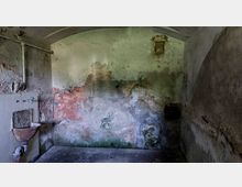 Ein alter, verlassener Raum mit stark beschädigten Wänden, teilweise abblätterndem Putz und Spuren von Feuchtigkeit. Links im Bild befindet sich ein rostiges Waschbecken mit veralteten Wasserrohren, während eine kleine, vergitterte Öffnung in der hinteren Wand sichtbar ist.