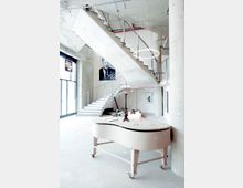 Heller, modern-industrieller Innenraum mit einer breiten Treppe aus Beton und Metallgeländer, einem weiß gestrichenen Flügel auf Rollen im Vordergrund und einer hängenden Lichtskulptur. Im Hintergrund befinden sich ein großes Fenster, ein Porträt an der Wand und mehrere Kerzen auf dem Klavier.