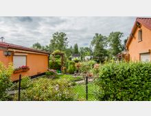 Zwei orangefarbene Wohnhäuser mit roten Dächern umgeben einen gepflegten Garten mit Blumen, Sträuchern und Rasenfläche, die durch einen niedrigen Drahtzaun begrenzt ist. Im Hintergrund sind weitere kleine Gebäude und hohe Bäume zu sehen, unter einem bewölkten Himmel.