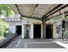 U-Bahn-Station mit offenen Bahnsteigen, einem überdachten Wartebereich und einem Schild mit der Aufschrift „Podbielskiallee“. Links führt ein Gleis in einen Tunnel, die Umgebung ist von Bäumen und einer Betonmauer umgeben.