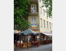 Straßenszene mit einer Pizzeria und einem angrenzenden Café, beide mit Außensitzbereichen mit Holztischen und -stühlen. Über einem der Eingänge hängt eine Regenbogenfahne mit der Aufschrift „PACE“, und grüne Bäume ragen in das Bild.
