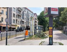 Bushaltestelle an einer Straße mit mehrstöckigen Wohnhäusern im Hintergrund, darunter Geschäfte im Erdgeschoss. Im Vordergrund stehen ein gelber Mülleimer, Fahrräder und ein Wartehäuschen, daneben führt ein Gehweg entlang einer Grünfläche mit Bäumen.