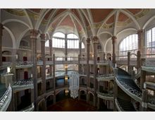 Großes, historisches Treppenhaus mit mehrstöckigen Galerien, verzierten Säulen und kunstvoll gestalteten schmiedeeisernen Geländern. Der Raum ist von hohen Fenstern mit Rundbögen und einem gewölbten, dekorierten Deckengewölbe mit goldenen Elementen geprägt.