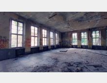 Verlassener Raum mit mehrfachen großen, rechteckigen Fenstern, die Licht einfallen lassen. Die Wände sind beschädigt, teilweise mit Graffiti bedeckt, und der Boden ist mit Schutt und heruntergefallenem Putz bedeckt.