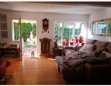 Ein Wohnzimmer mit hellbraunem Parkettboden und Tageslicht, das durch große Fenster und eine geöffnete Terrassentür einfällt. Rechts steht eine großzügige Sofalandschaft mit gemusterten Decken und Kissen; auf der Fensterbank stehen zahlreiche Topfpflanzen, darunter Kakteen. Links führt die Tür zu einer Terrasse mit Gartenmöbeln und einer weißen Statue im Hintergrund. Ein Hund steht vor der geöffneten Tür.