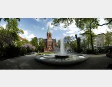 Ein Park mit einem großen Springbrunnen im Vordergrund, umgeben von Bäumen und Sitzbänken. Im Hintergrund steht eine rote Backsteinkirche mit spitzem grünen Turm, flankiert von Wohngebäuden.