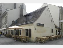 Historisches Gebäude mit gelber Fassade und Ziegeldach, beschriftet als „Weinhaus“, mit einer Außengastronomie, die mehrere Holztische und Stühle unter Sonnenschirmen umfasst. Das Gebäude steht in einer städtischen Umgebung, angrenzend an andere ältere Bauwerke.