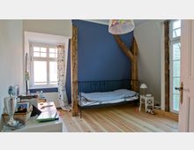 Schlafzimmer mit blauer Wand, freiliegenden Holzbalken und einem Einzelbett mit Metallrahmen. Holzdielenboden, ein Nachttisch mit Lampe neben dem Bett und ein Fenster mit weißen Vorhängen versehen mit blauem Blumendruck lassen viel Tageslicht herein.