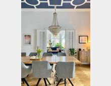 Esszimmer mit modernem, hellem Holztisch und grauen gepolsterten Stühlen, darunter ein Fischgrätenparkettboden. An der Decke hängt ein großer Kristallleuchter, und durch geöffnete Doppeltüren ist ein Wohnzimmer mit Pflanzen und einem Sofa sichtbar.