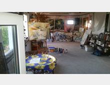 Ein Atelierraum mit mehreren abstrakten Gemälden, die an den Wänden und auf Staffeleien stehen. Im Vordergrund befindet sich ein Tisch mit einem blau-gelben Tischtuch und Malutensilien, im Hintergrund sind Regale mit Büchern und Malwerkzeugen sichtbar.