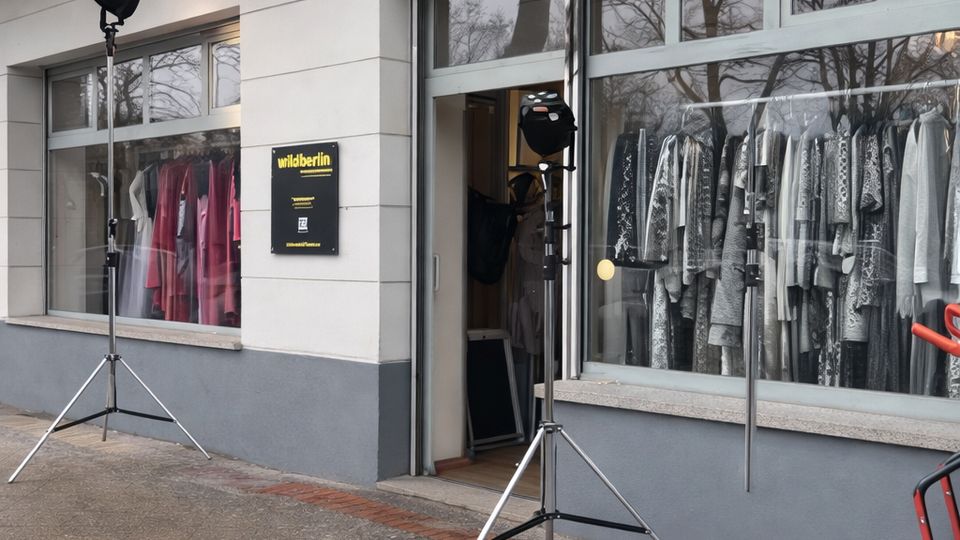 Schaufensterfront eines Modegeschäfts in einem hellen Gebäude mit großem gelben „wildberlin“-Schild über der Eingangstür. Große Fenster zeigen Kleider auf Bügeln, davor stehen zwei Beleuchtungsstative auf dem Gehweg.