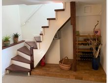 Treppenhaus mit einer nach oben führenden Holztreppe, weißen Wänden und einem kleinen dekorativen Bereich darunter. Rechts befindet sich ein Weinregal, daneben ein Korb und ein blauer Topf mit Trockenästen, während links Zimmerpflanzen auf einem breiten Holzgeländer stehen.