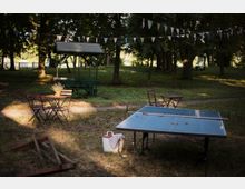 Ein ruhiger Außenbereich in einem kleinen Park mit Tischen und Stühlen aus Holz sowie einer Tischtennisplatte. Im Hintergrund ist ein hölzerner Pavillon und eine leuchtend grüne Wiese von Bäumen umgeben; bunte Wimpel hängen zwischen den Bäumen.