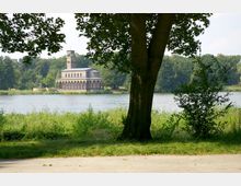 Ein historisches Kirchengebäude mit Arkaden und einem Glockenturm am Ufer eines Sees, umgeben von dichtem Wald. Im Vordergrund sind ein Baum und Grasflächen zu sehen, die den See einrahmen.