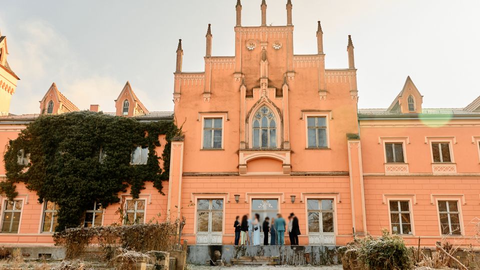 Rosafarbenes, schlossähnliches Gebäude im neugotischen Stil mit zentralem Giebel, schlanken Ziertürmchen und Spitzbogenfenster über dem Eingang. Links bedeckt dichter Efeu einen großen Teil der Fassade; davor liegt ein Gartenbereich mit Beeten und Wegen.