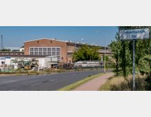 Industriegelände mit einem großen Backsteingebäude, Lagerflächen und Baumaschinen. Im Vordergrund eine asphaltierte Straße, ein Gehweg und ein Schild mit der Aufschrift „Industriepark Ost“.