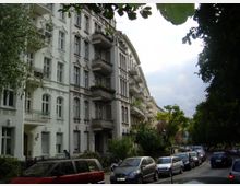 Straßenansicht mit historischen Mehrfamilienhäusern im wilhelminischen Stil, gekennzeichnet durch verzierte Fassaden, Balkone und große Fensterelemente; geparkte Autos säumen die Straße, die von Bäumen und Grünflächen gesäumt ist.