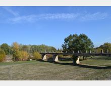 Betonbrücke mit abgeschnittenen Geländerpfosten in einer flachen Graslandschaft, umgeben von Büschen und Bäumen mit herbstlichem Laub; im Hintergrund ein klarer blauer Himmel.