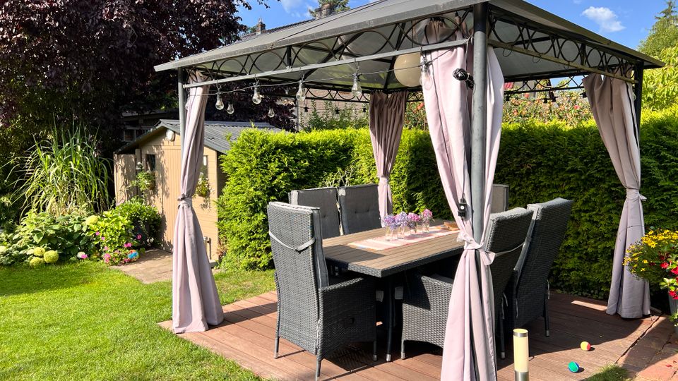 Überdachte Gartensitzecke mit Metallpavillon und seitlichen Vorhängen auf einer Holzterrasse, darunter ein Esstisch mit mehreren geflochtenen Stühlen. Umgeben von Rasen, hohen Hecken, Blumenbeeten und einem kleinen Gartenhaus im Hintergrund.