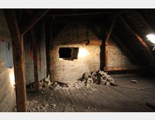 Ein leerer Dachboden mit Holzbalken und einer Ziegelwand, in der ein großes Loch sichtbar ist. Auf dem Boden liegen Schutthaufen und zerbrochene Ziegel, während spärliche Beleuchtung den Raum erhellt.
